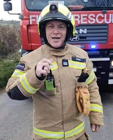 A smiling Dave Evans, wearing fire kit, standing in front of a fire engine